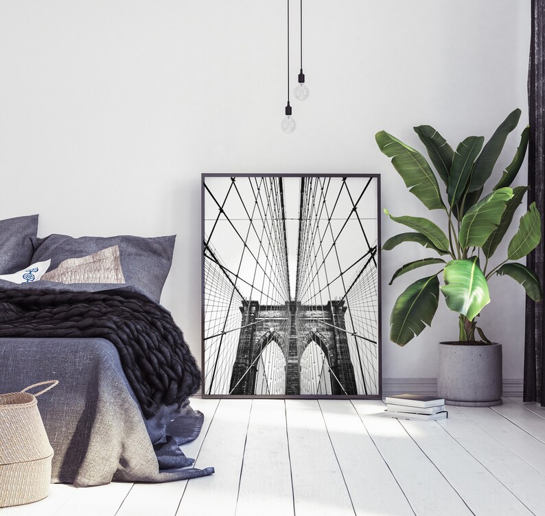 Pu&ograve; includere: Stampa incorniciata in bianco e nero del ponte di Brooklyn. L'opera d'arte &egrave; esposta in una camera da letto moderna con biancheria da letto grigia, un cesto di vimini e una pianta in vaso. L'ambiente &egrave; minimalista.