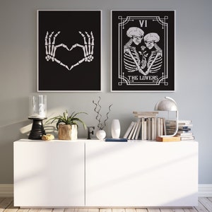 May include: Two black and white framed prints with skeleton imagery. The left print shows two skeletal hands forming a heart shape. The right print shows two skeletons embracing with a rose between them. The text "VI THE LOVERS" is below the skeletons. The prints are displayed on a white shelf with a lamp, books, and other decor.