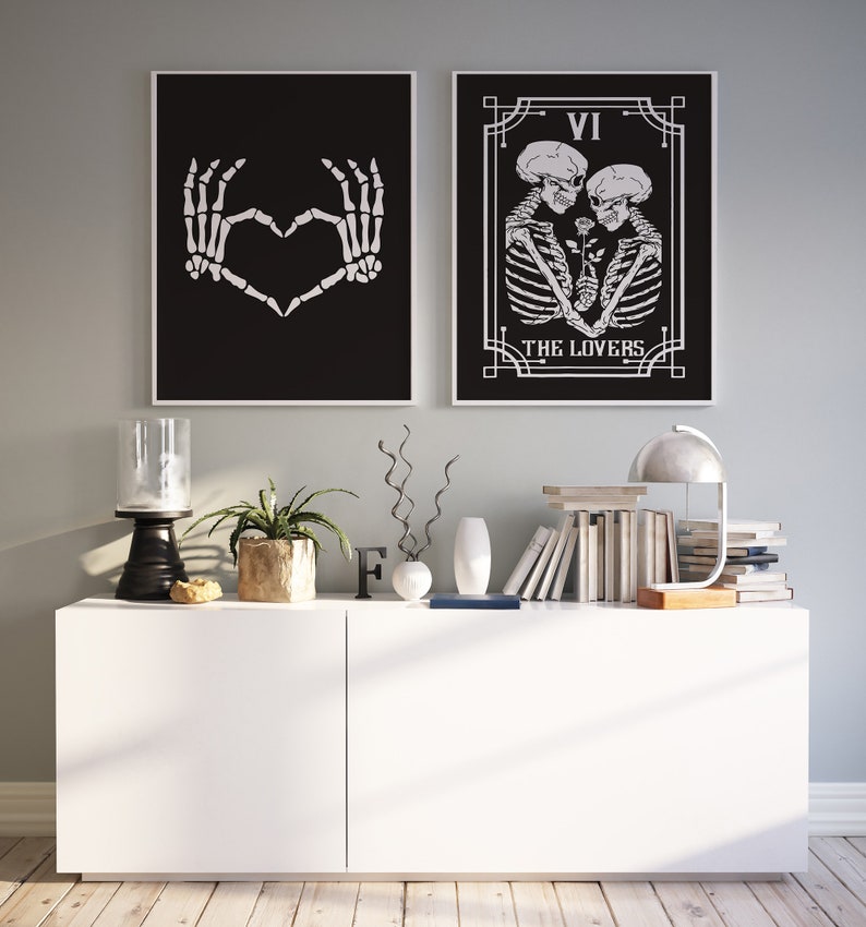 May include: Two black and white framed prints with skeleton imagery. The left print shows two skeletal hands forming a heart shape. The right print shows two skeletons embracing with a rose between them. The text "VI THE LOVERS" is below the skeletons. The prints are displayed on a white shelf with a lamp, books, and other decor.