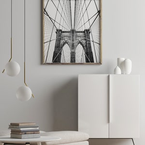Pu&ograve; includere: Fotografia in bianco e nero del ponte di Brooklyn, incorniciata in legno chiaro. L'immagine cattura l'intricata struttura dei cavi e le torri in pietra del ponte, creando un'impressionante esposizione architettonica. L'opera d'arte &egrave; esposta in un interno moderno.