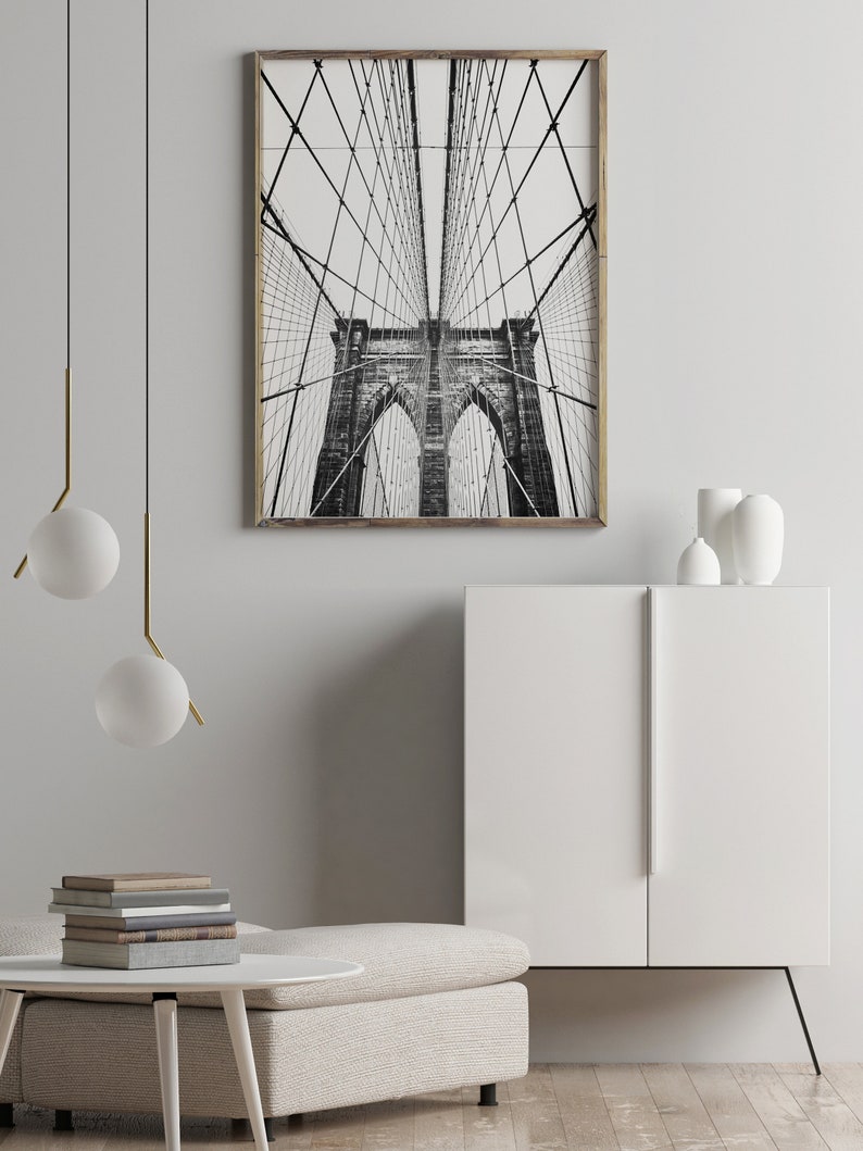 Pu&ograve; includere: Fotografia in bianco e nero del ponte di Brooklyn, incorniciata in legno chiaro. L'immagine cattura l'intricata struttura dei cavi e le torri in pietra del ponte, creando un'impressionante esposizione architettonica. L'opera d'arte &egrave; esposta in un interno moderno.