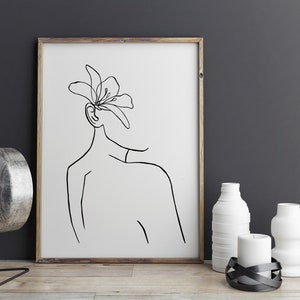 May include: A black and white line drawing of a person with a flower on their head. The drawing is framed in a wooden frame and is hanging on a gray wall.