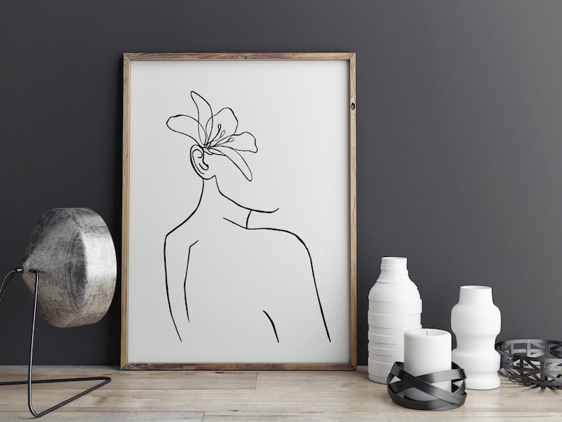 May include: A black and white line drawing of a person with a flower on their head. The drawing is framed in a wooden frame and is hanging on a gray wall.