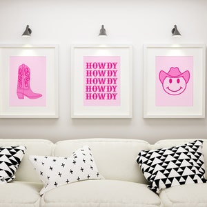 May include: Three framed prints with pink backgrounds and pink outlines. The left print features a pink cowboy boot. The middle print has the word "HOWDY" repeated five times. The right print features a pink smiley face wearing a cowboy hat.