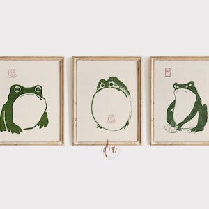 May include: Three framed prints of green frogs in different poses. The frogs are stylized and have a simple, minimalist design. The prints are set against a white background.