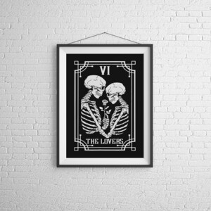 May include: Black and white illustration of the Lovers tarot card, featuring two skeletons holding a rose. The card is framed in a black frame with a white mat. The card number VI and the title "THE LOVERS" are printed on the card.