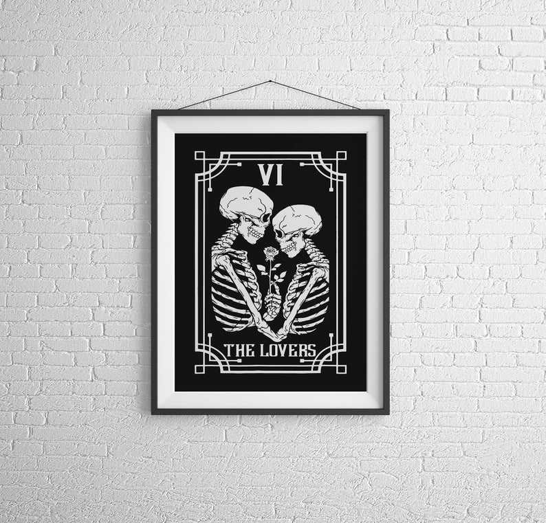 May include: Black and white illustration of the Lovers tarot card, featuring two skeletons holding a rose. The card is framed in a black frame with a white mat. The card number VI and the title "THE LOVERS" are printed on the card.