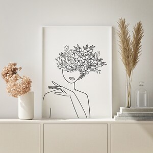 May include: Line art print of a woman's face with flowers growing from her head. The print is in black and white and is framed in a white frame.
