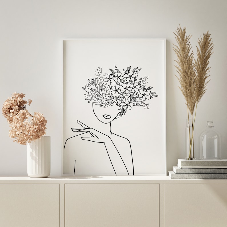 May include: Line art print of a woman's face with flowers growing from her head. The print is in black and white and is framed in a white frame.