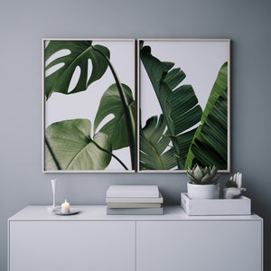 May include: Two framed prints of tropical plants with green leaves against a white background. The prints are hanging on a light grey wall above a white dresser with a small candle and a potted succulent.