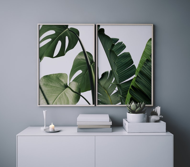 May include: Two framed prints of tropical plants with green leaves against a white background. The prints are hanging on a light grey wall above a white dresser with a small candle and a potted succulent.
