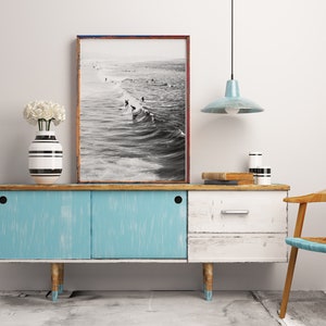 May include: A blue and white vintage sideboard with a black and white striped vase with white flowers on top. A black and white photograph of surfers in the ocean hangs above the sideboard. A light blue pendant light hangs from the ceiling above the sideboard. A wooden chair with a woven seat sits to the right of the sideboard.