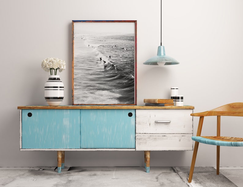 May include: A blue and white vintage sideboard with a black and white striped vase with white flowers on top. A black and white photograph of surfers in the ocean hangs above the sideboard. A light blue pendant light hangs from the ceiling above the sideboard. A wooden chair with a woven seat sits to the right of the sideboard.