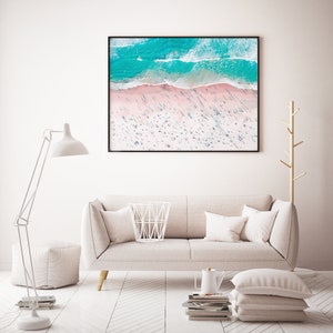Op de afbeelding: Een witte bank met kussens staat in een woonkamer met een witte vloer en een witte lamp. Een ingelijst foto van een strandsc&egrave;ne hangt aan de muur boven de bank.