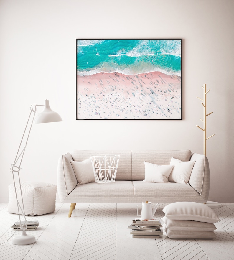 Op de afbeelding: Een witte bank met kussens staat in een woonkamer met een witte vloer en een witte lamp. Een ingelijst foto van een strandsc&egrave;ne hangt aan de muur boven de bank.