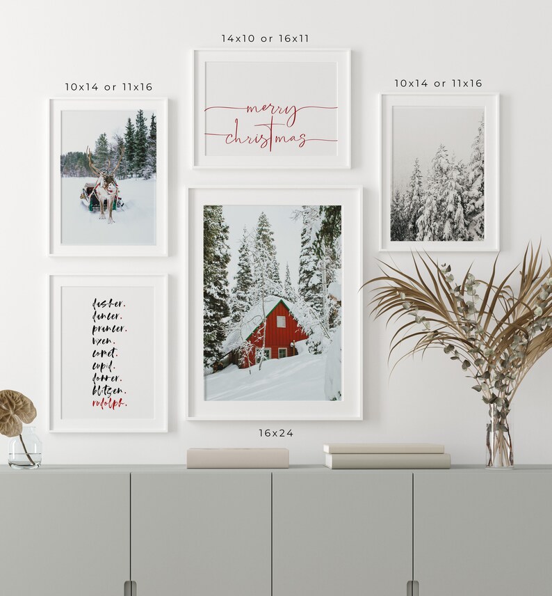 Op de afbeelding: Een set van vier witte ingelijste prints met winterse taferelen. De bovenste print zegt "vrolijk kerstfeest" in rode cursieve letters. De linker print heeft een zwart-wit foto van een rendier dat een slee trekt. De rechter print heeft een zwart-wit foto van een besneeuwd bos. De onderste print heeft een zwart-wit foto van een rood huisje in een besneeuwd bos. De prints zijn gerangschikt op een witte muur boven een grijze kast.