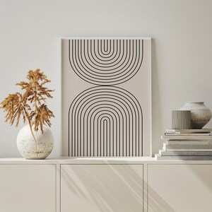 May include: A framed print with a minimalist design of black arches on a light beige background. The print is displayed on a white shelf with a vase of dried flowers and a stack of books.