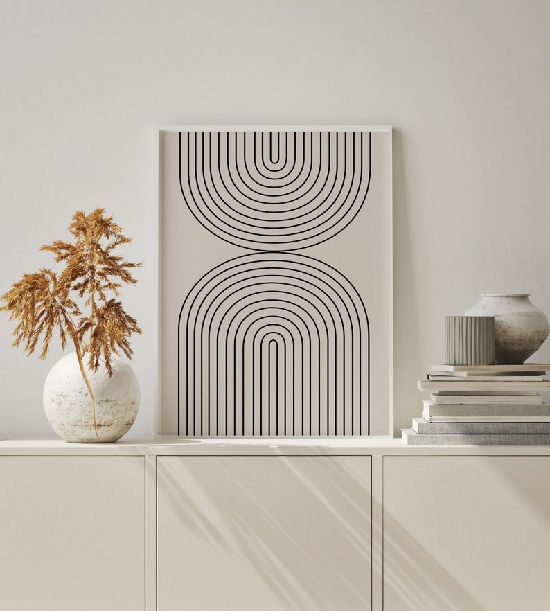 May include: A framed print with a minimalist design of black arches on a light beige background. The print is displayed on a white shelf with a vase of dried flowers and a stack of books.