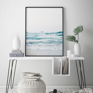 Peut inclure: Un bureau blanc avec des pieds en &eacute;pingle &agrave; cheveux noirs et une &eacute;tag&egrave;re. Une photographie encadr&eacute;e d'un oc&eacute;an bleu avec du sable blanc est au-dessus du bureau. Un objet d&eacute;coratif en forme d'ananas blanc est sur le bureau. Un pouf blanc avec une texture tiss&eacute;e est devant le bureau.
