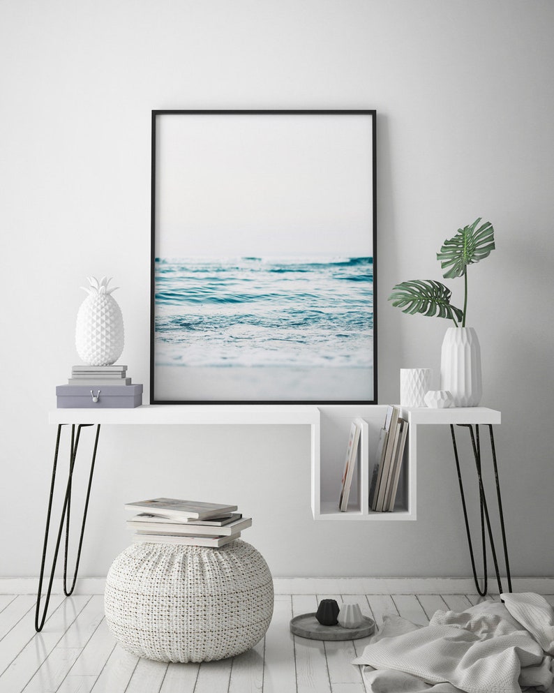 Peut inclure: Un bureau blanc avec des pieds en &eacute;pingle &agrave; cheveux noirs et une &eacute;tag&egrave;re. Une photographie encadr&eacute;e d'un oc&eacute;an bleu avec du sable blanc est au-dessus du bureau. Un objet d&eacute;coratif en forme d'ananas blanc est sur le bureau. Un pouf blanc avec une texture tiss&eacute;e est devant le bureau.