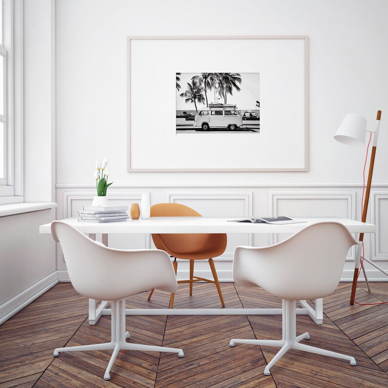 May include: A black and white photograph of a vintage van with surfboards on the roof, parked on a beach with palm trees in the background. The photo is framed and hanging on a white wall above a white table with two white chairs and a brown chair.