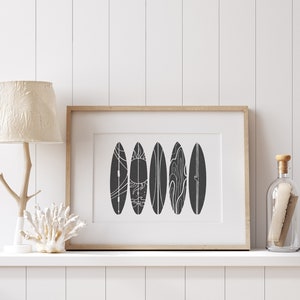 May include: A framed black and white print of five stylized surfboards with white line designs. The print is hanging on a white wall above a shelf with a lamp and a decorative coral.