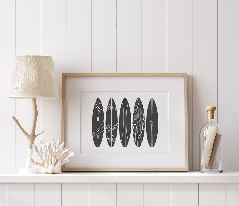 May include: A framed black and white print of five stylized surfboards with white line designs. The print is hanging on a white wall above a shelf with a lamp and a decorative coral.