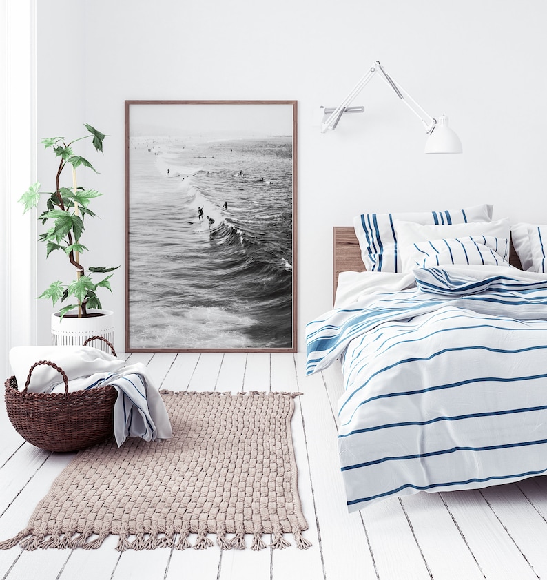 May include: A white bedroom with a bed with blue and white striped bedding. A black and white photograph of surfers hangs on the wall above the bed. A woven rug is on the floor in front of the bed.
