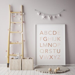 May include: A white framed poster with the alphabet in brown letters on a white background. The poster is hanging on a light pink wall with a string of white and pink pom poms hanging on a wooden ladder to the left of the poster. There is a beige rug with a circle pattern on the floor in front of the poster.