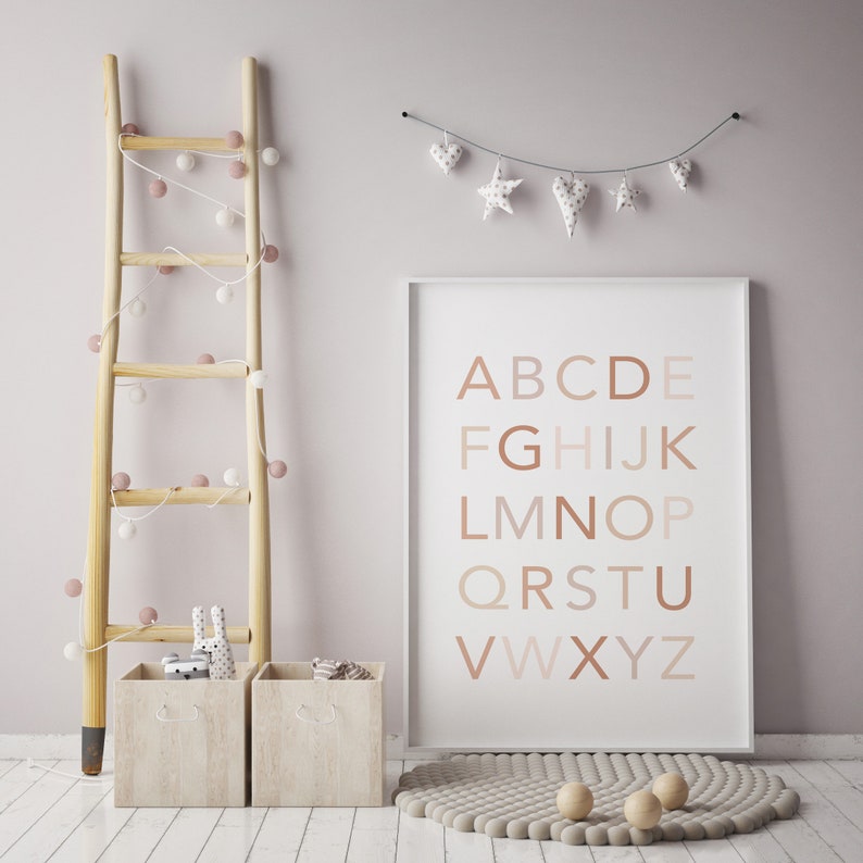May include: A white framed poster with the alphabet in brown letters on a white background. The poster is hanging on a light pink wall with a string of white and pink pom poms hanging on a wooden ladder to the left of the poster. There is a beige rug with a circle pattern on the floor in front of the poster.