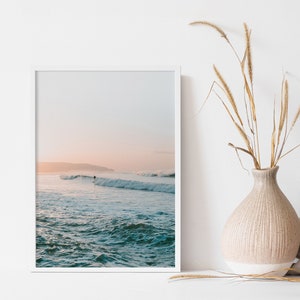 May include: A framed photograph of a surfer riding a wave in the ocean at sunset. The sky is a soft pink and blue, and the water is a deep blue.
