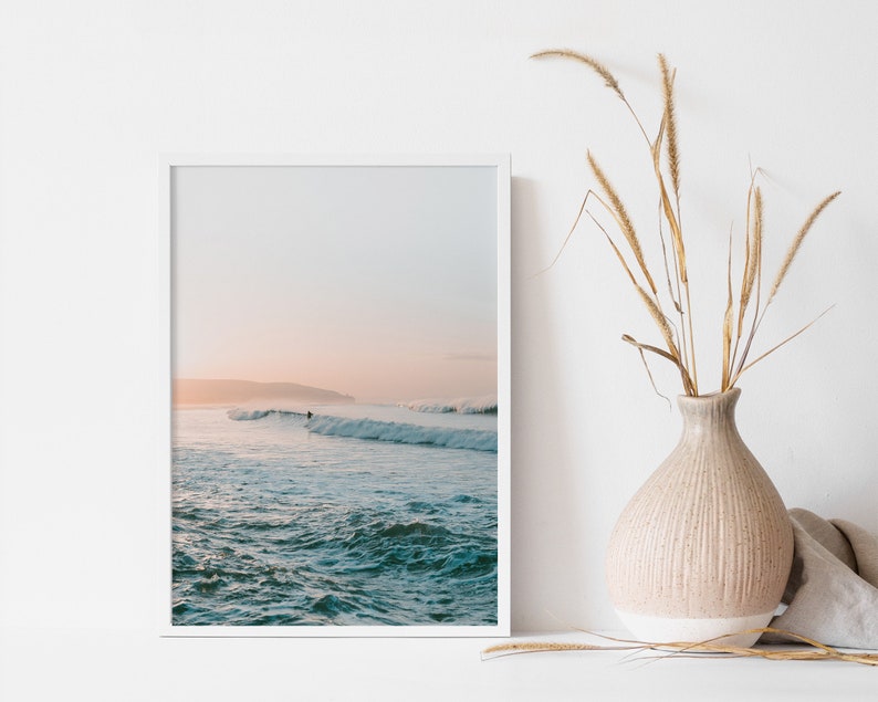 May include: A framed photograph of a surfer riding a wave in the ocean at sunset. The sky is a soft pink and blue, and the water is a deep blue.