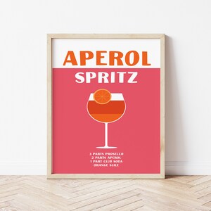 Pode incluir: Um cartaz rosa e branco com o texto "Aperol Spritz" em laranja e branco. O cartaz apresenta um copo de Aperol Spritz com uma fatia de laranja no topo. Os ingredientes s&atilde;o listados abaixo do copo: 3 partes de Prosecco, 2 partes de Aperol, 1 parte de soda club, fatia de laranja.