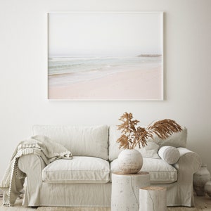 May include: A white sofa with a throw blanket, a vase with dried grasses, and two white wooden stumps as side tables. A framed photograph of a beach scene hangs on the wall behind the sofa.