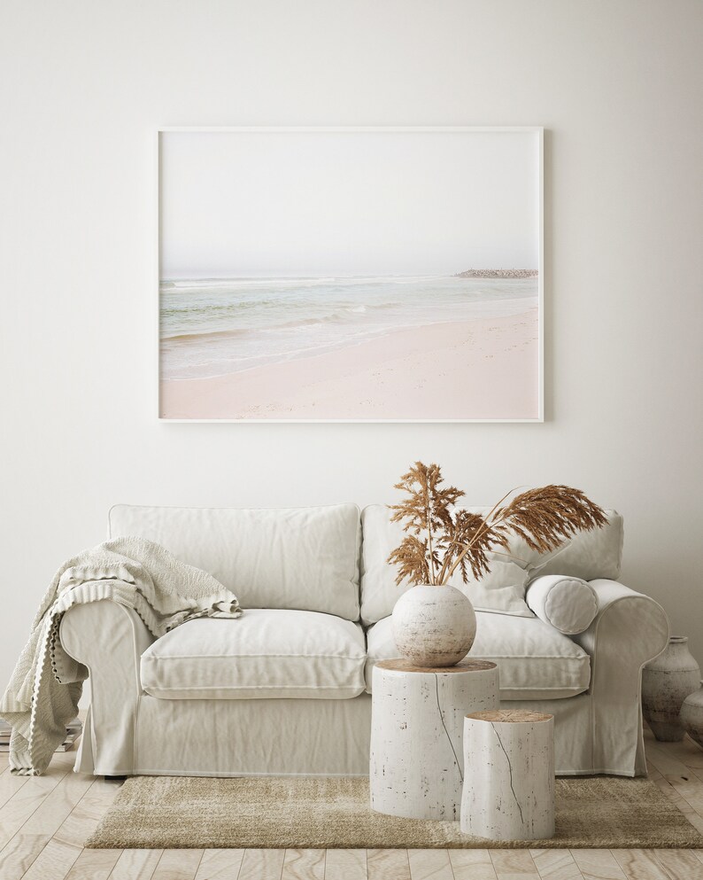 May include: A white sofa with a throw blanket, a vase with dried grasses, and two white wooden stumps as side tables. A framed photograph of a beach scene hangs on the wall behind the sofa.