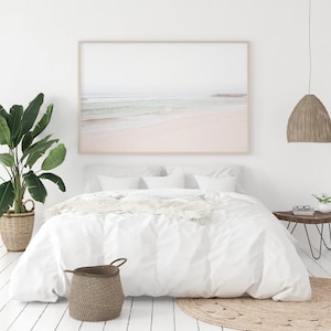 May include: A white bed with white bedding and a white throw blanket. A large framed photograph of a beach scene hangs above the bed. A potted plant is on the left side of the bed and a small side table with a plant and glasses is on the right side of the bed. A woven rug is on the floor in front of the bed.
