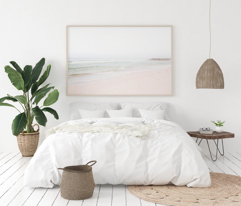 May include: A white bed with white bedding and a white throw blanket. A large framed photograph of a beach scene hangs above the bed. A potted plant is on the left side of the bed and a small side table with a plant and glasses is on the right side of the bed. A woven rug is on the floor in front of the bed.