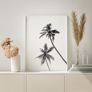 May include: Black and white print of three palm trees against a white background. The print is framed in a white frame and is hanging on a white wall.