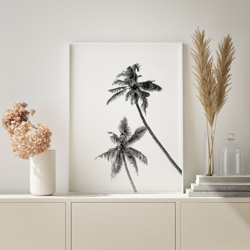 May include: Black and white print of three palm trees against a white background. The print is framed in a white frame and is hanging on a white wall.