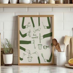 May include: A framed print with a white background and green illustrations of hands holding various kitchen utensils. The text "bon appetit" is written in the centre of the print.