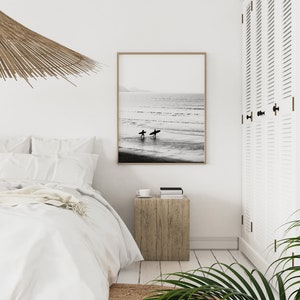 May include: Black and white photograph of two surfers walking on a beach with their surfboards. The photo is framed and hanging on a white wall in a bedroom.