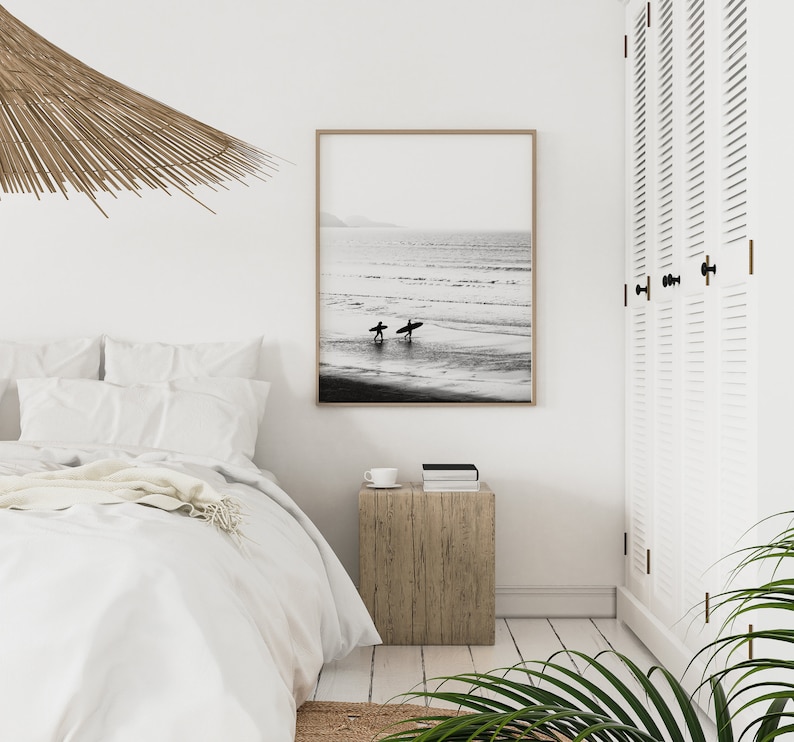 May include: Black and white photograph of two surfers walking on a beach with their surfboards. The photo is framed and hanging on a white wall in a bedroom.