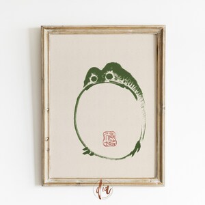 May include: A framed print of a green circle with a red stamp in the center. The circle is a stylized image of a frog. The print is on a beige background.