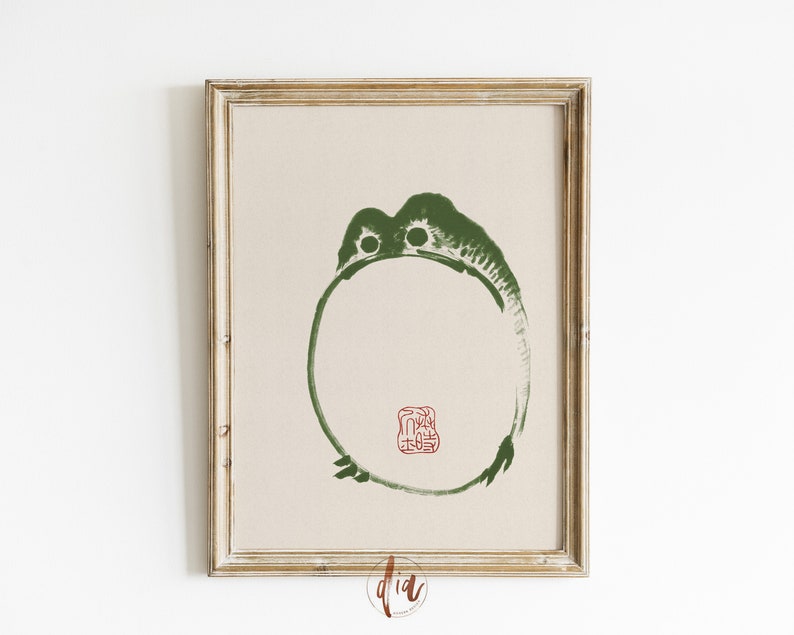 May include: A framed print of a green circle with a red stamp in the center. The circle is a stylized image of a frog. The print is on a beige background.