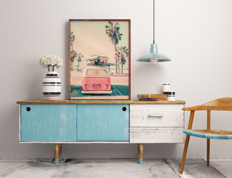 May include: A vintage pink Volkswagen van with surfboards on the roof parked in front of a beach scene with palm trees. The van has a license plate that says "SURF'S UP". The image is printed on a canvas and framed in a wooden frame.
