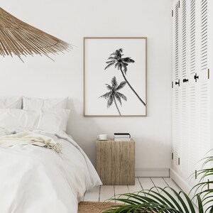 May include: Black and white photograph of palm trees against a white background, framed in a light wood frame. The photograph is hanging on a white wall in a bedroom setting.