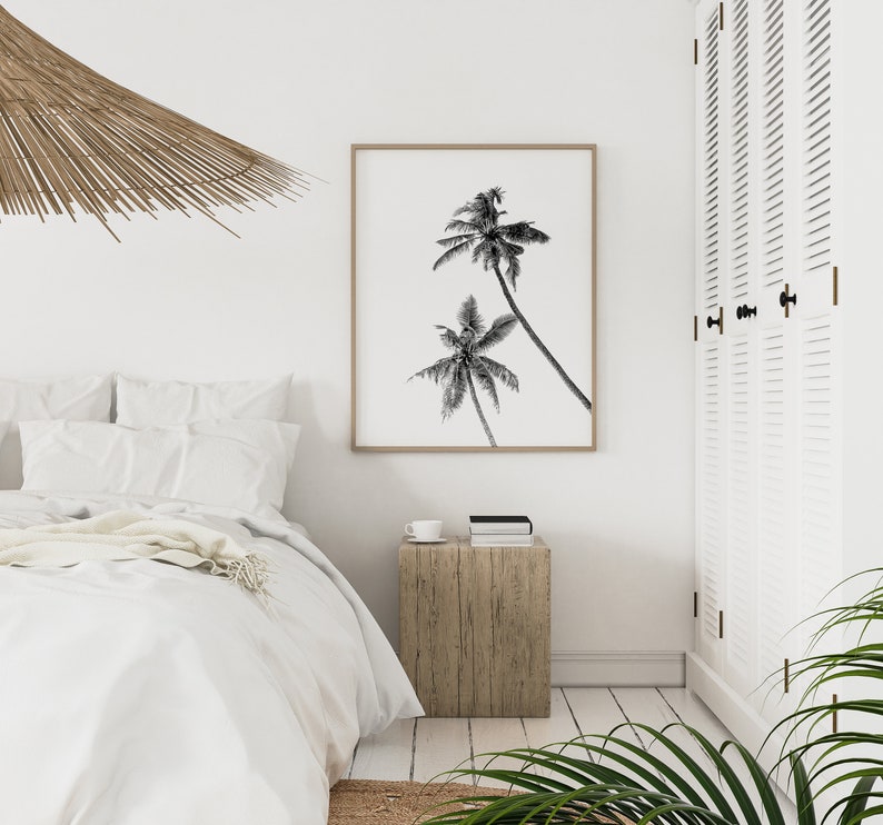 May include: Black and white photograph of palm trees against a white background, framed in a light wood frame. The photograph is hanging on a white wall in a bedroom setting.