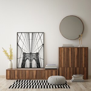 Pu&ograve; includere: Una stampa in bianco e nero incorniciata del ponte di Brooklyn &egrave; appesa sopra un mobile basso in legno. Uno specchio rotondo con cornice in bronzo &egrave; montato a parete. Un tappeto a strisce bianche e nere e un pouf grigio sono sul pavimento.