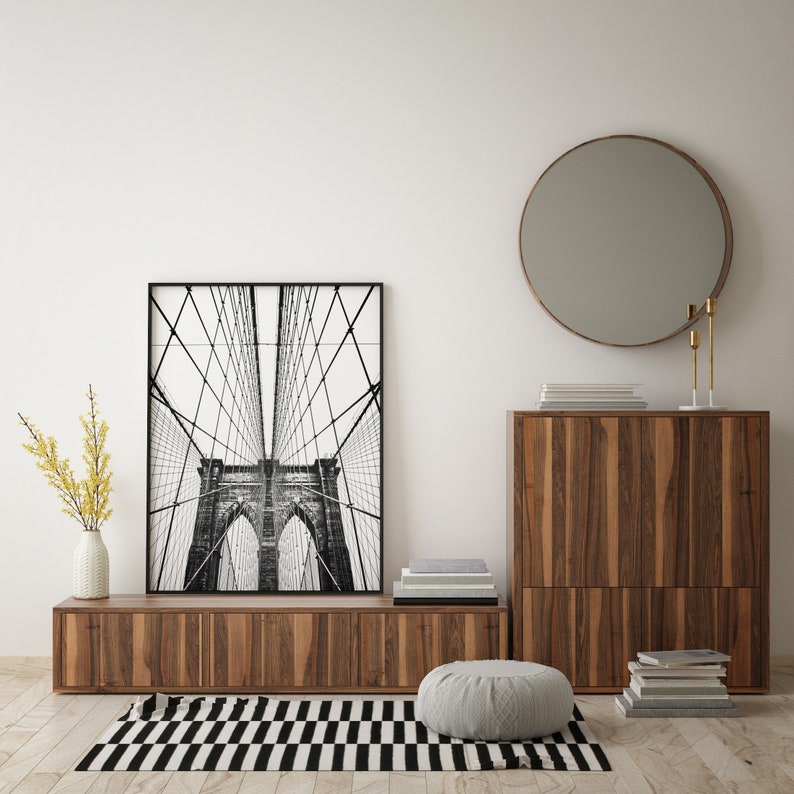 Pu&ograve; includere: Una stampa in bianco e nero incorniciata del ponte di Brooklyn &egrave; appesa sopra un mobile basso in legno. Uno specchio rotondo con cornice in bronzo &egrave; montato a parete. Un tappeto a strisce bianche e nere e un pouf grigio sono sul pavimento.
