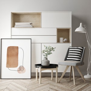 Op de afbeelding: Een witte kast met een zwart ingelijst abstract kunstwerk, een kleine ronde houten tafel met een zwarte dienblad, een vaas met groene bladeren en een grijze stoel met een zwart-wit gestreept kussen. De kast heeft een plank met boeken en een klein open vak met een witte vaas en een klein wit voorwerp.
